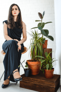 Portrait of jazz vocalist Issey Chivers seated beside potted plants against a white brick wall.